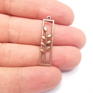 May include: A copper-colored metal pendant with a rectangular frame and a stylized leaf design inside. The pendant has a loop at the top for attaching to a chain or cord.