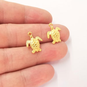 May include: Two small, gold-coloured turtle charms. Each charm has a loop at the top for hanging and detailed turtle features. The charms are displayed against a light background.