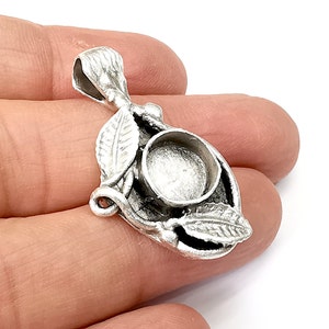 May include: Silver pendant with a round bezel for a cabochon stone. The pendant is decorated with leaves and vines.