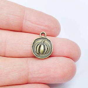 May include: A small, antique brass-coloured pumpkin charm. The charm is round with a raised pumpkin design in the centre, surrounded by a beaded border. It has a small loop at the top for attaching to jewellery or other crafts.