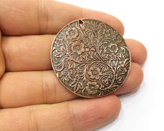 Op de afbeelding: Een ronde, koperkleurige metalen hanger met een reliëf bloemenpatroon. De hanger heeft een gedetailleerd ontwerp van bloemen, bladeren en wijnranken. Het heeft een klein gaatje aan de bovenkant om op te hangen.