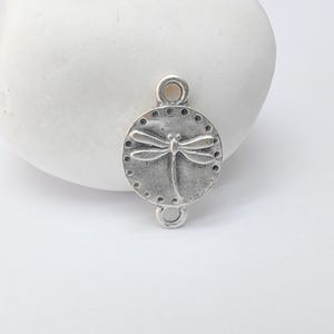 May include: A silver-toned metal charm featuring a dragonfly design. The charm is circular with a dragonfly in the center and small dot details around the edge. It has loops at the top and bottom for attachment.