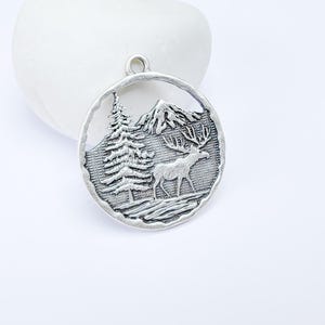 Puede incluir: Encanto de plata con un diseño detallado de una montaña, un pino y un ciervo. El encanto es redondo y tiene una superficie texturizada.