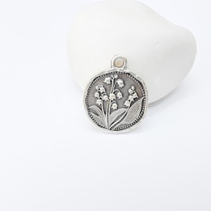May include: A round, silver-toned pendant featuring a detailed relief of lily of the valley flowers and leaves. The pendant has a textured border and a small loop at the top for a chain. The design is set against a white background.