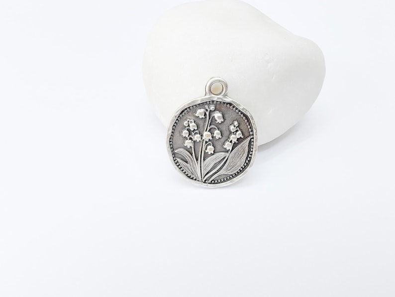 May include: A round, silver-toned pendant featuring a detailed relief of lily of the valley flowers and leaves. The pendant has a textured border and a small loop at the top for a chain. The design is set against a white background.