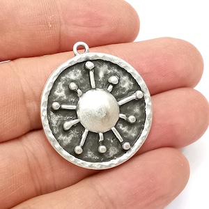 May include: Silver pendant with a circular design featuring a sunburst with a textured surface.
