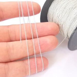 May include: A spool of silver chain, approximately 1.5 mm in width, is shown on a white background. The chain is being held by a hand, and the spool is visible in the background.