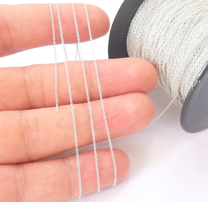 May include: A spool of silver chain, approximately 1.5 mm in width, is shown on a white background. The chain is being held by a hand, and the spool is visible in the background.