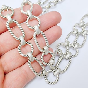 May include: Silver-toned necklace featuring a chunky chain design. The necklace has oval-shaped links with a twisted rope texture, connected by smooth, cylindrical bars. The jewelry is displayed against a light background.