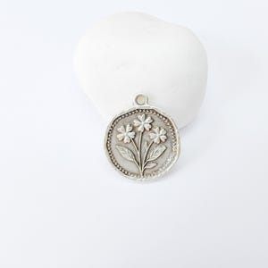 May include: A silver-colored pendant with a floral design. The pendant features three flowers with leaves and a beaded border. The pendant is round and has a small loop at the top for a chain. The background is white.