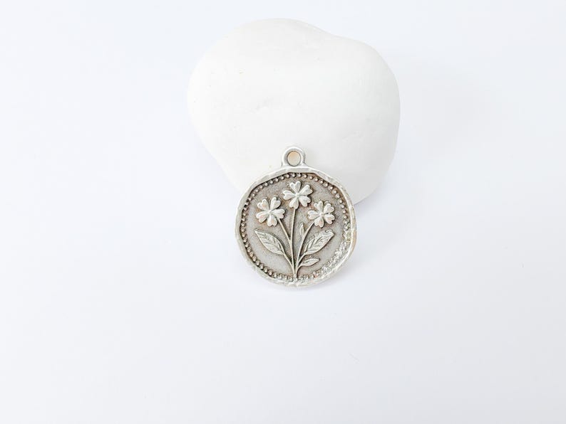 May include: A silver-colored pendant with a floral design. The pendant features three flowers with leaves and a beaded border. The pendant is round and has a small loop at the top for a chain. The background is white.