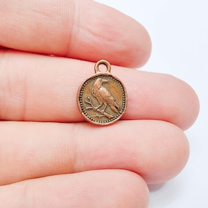 Pode incluir: Um pequeno pingente de metal em tom de cobre com um design em relevo de um pássaro pousado num galho. O pingente circular apresenta uma borda com contas e uma pequena argola no topo, com um acabamento envelhecido de estilo antigo.