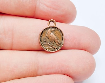 May include: A small, round, copper-toned metal charm featuring a raised relief design of a bird perched on a branch. The circular charm has a beaded border and a small loop at the top for attachment, showcasing a weathered, antique-style finish.