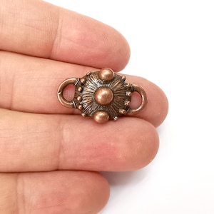 Puede incluir: Un cierre de metal de color cobre con un diseño circular y dos bucles. El cierre tiene una superficie texturizada y un centro elevado.