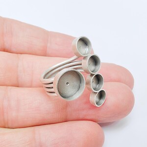 May include: A silver-toned ring with multiple circular settings of varying sizes. The ring features a coiled band and several cup-like holders, designed to hold small items. The ring is held in a hand against a white background.