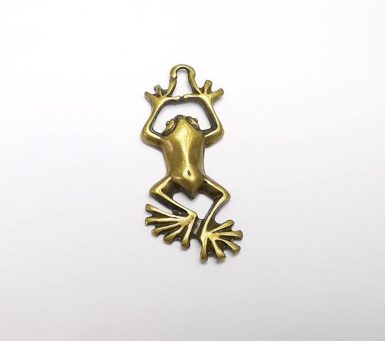 May include: A gold-toned metal frog charm with a loop at the top for hanging. The frog is in a sitting position with its legs outstretched.