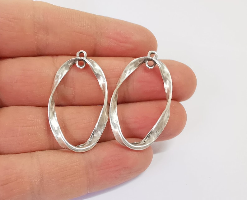 May include: Two silver oval-shaped pendant charms with a twisted design. The charms have a loop at the top for attaching to a necklace or bracelet.