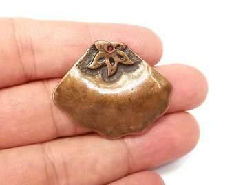 May include: A bronze-colored pendant with a scalloped edge and a stylized flower design. The flower has a red center and is set against a textured background.