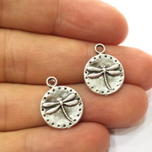 May include: Two silver-toned metal charms in the shape of a circle with a dragonfly design. The dragonfly is raised and has a detailed design.