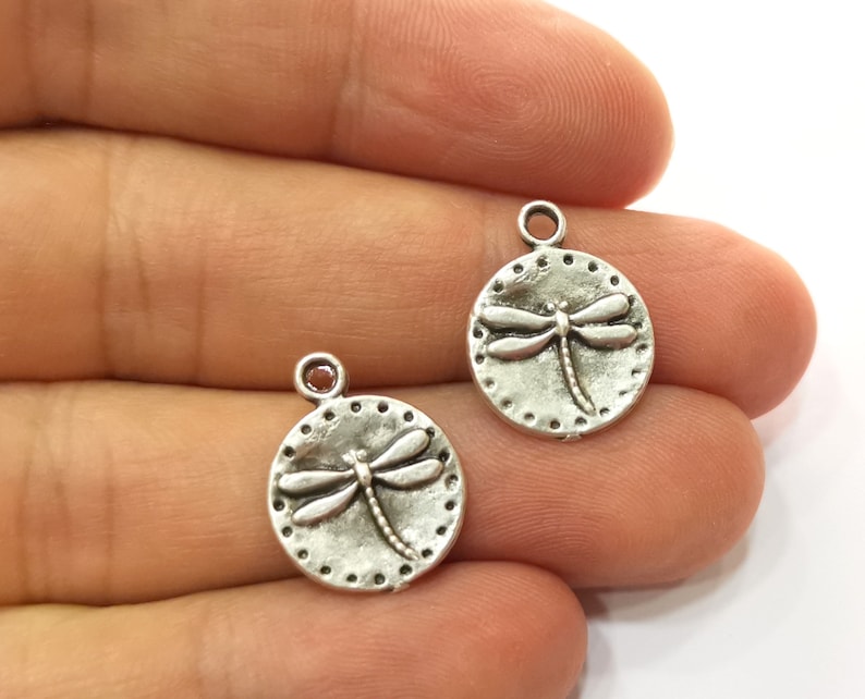 May include: Two silver-toned metal charms in the shape of a circle with a dragonfly design. The dragonfly is raised and has a detailed design.