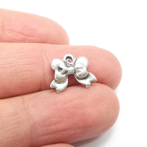 Könnte beinhalten: Silberner Anhänger in Form einer Schleife mit einem roten Strassstein in der Mitte. Der Anhänger hat oben ein Loch zum Anbringen an einem Armband oder einer Halskette.