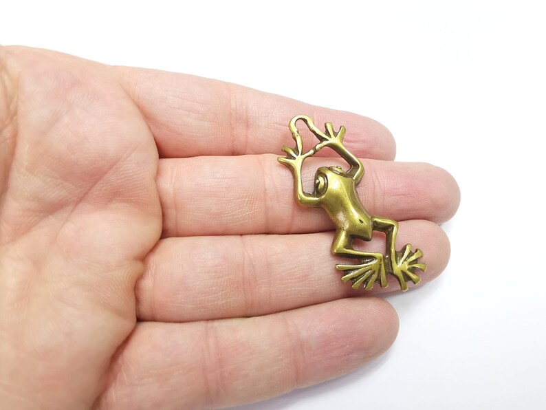 May include: A gold-toned metal frog charm with a detailed design. The frog is in a climbing pose with its legs outstretched.
