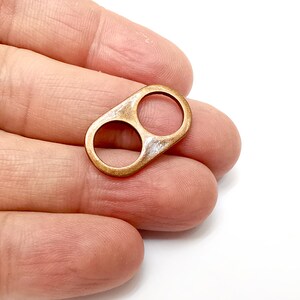 Op de afbeelding: Een koperkleurige metalen bedel met twee cirkelvormige openingen en een rechthoekige vorm. De bedel wordt in een hand gehouden tegen een witte achtergrond. De bedel is waarschijnlijk voor het maken van sieraden of knutselen.