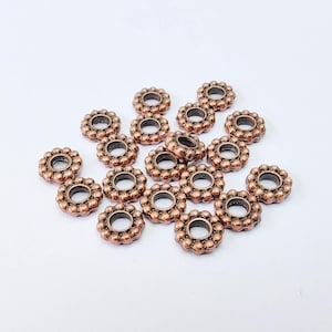 Peut inclure: Un ensemble de perles en forme de fleur, de couleur cuivrée. Chaque perle présente un trou central entouré d'un anneau de petits points en relief. Les perles sont disposées sur une surface blanche, parfaites pour la création de bijoux ou l'artisanat.