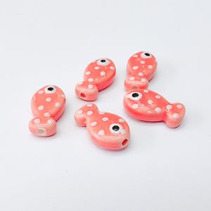 May include: A collection of six coral-colored ceramic fish beads with white polka dots. Each bead has a black eye and a hole for stringing. These beads are ideal for jewelry making and craft projects.
