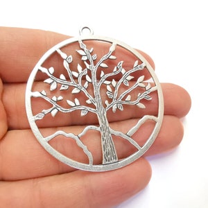 May include: Silver-toned metal charm in the shape of a tree of life. The charm is circular and features a detailed design of a tree with branches and leaves.