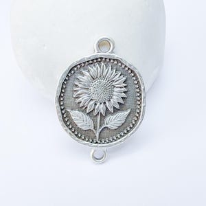 May include: A silver-toned metal charm featuring a detailed sunflower design. The charm has a textured background and a decorative border with two loops for attachment. The sunflower has detailed petals and leaves.