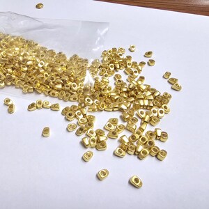 May include: A collection of small, gold-colored metal beads, scattered on a white surface. The beads are rectangular with a hole in the center, and some are spilling from a clear plastic bag. The lighting is bright and the background is plain.