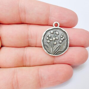 May include: A silver-colored round charm with a detailed relief of lily of the valley flowers and leaves. The charm has a small loop at the top for attaching to a chain or bracelet. The edges have a textured, beaded design.