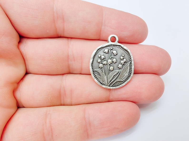 May include: A silver-colored round charm with a detailed relief of lily of the valley flowers and leaves. The charm has a small loop at the top for attaching to a chain or bracelet. The edges have a textured, beaded design.