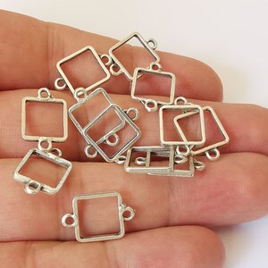 May include: A selection of small, silver-coloured square connector charms. Each charm has a square frame with loops on each side for attachment. The charms are held in a hand against a white backdrop.