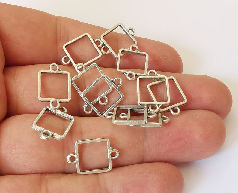 May include: A selection of small, silver-coloured square connector charms. Each charm has a square frame with loops on each side for attachment. The charms are held in a hand against a white backdrop.