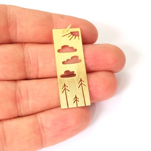 Puede incluir: Encanto de metal dorado con un diseño recortado de un sol, nubes y árboles. El encanto es rectangular y tiene un bucle en la parte superior para sujetarlo a un collar o pulsera.
