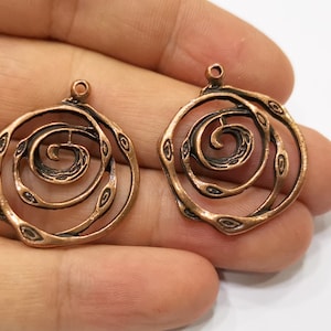 May include: Two copper-coloured spiral-shaped pendants. Each pendant features a central spiral design surrounded by a textured, openwork frame. The pendants have a small loop at the top for attaching to a chain or other jewellery component.