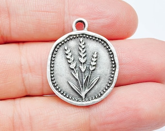 Puede incluir: Un dije redondo plateado con un diseño en relieve de tres ramitas de lavanda. El dije tiene un borde con cuentas y un pequeño lazo en la parte superior para sujetarlo a una joya. El dije está en la mano de una persona.