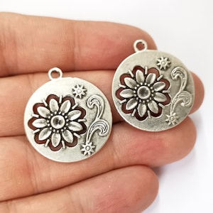 Op de afbeelding: Twee zilverkleurige metalen bedels met een bloemmotief. Elke bedel heeft een grote bloem met een kleinere bloem in het midden, omringd door ingewikkelde krulwerk.