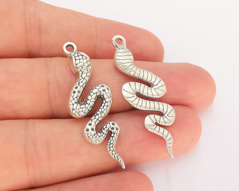 May include: Two silver snake charms with detailed scales. The charms are in the shape of a snake curled up with its tail in its mouth.