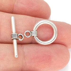 May include: Silver toggle clasp with a round ring and a bar with a decorative twist. The clasp is a jewellery finding used for necklaces, bracelets, and other jewellery.