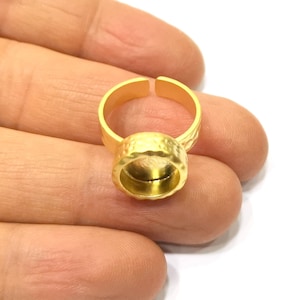 May include: A gold-colored adjustable ring with a textured bezel setting. The ring's band is open, allowing for size adjustment. The bezel setting is designed to hold a gemstone or other decorative element. The ring is shown against a neutral background.