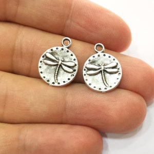 May include: Two silver-toned metal charms in the shape of a circle with a dragonfly design. The dragonfly is embossed on the charm and has a detailed design.