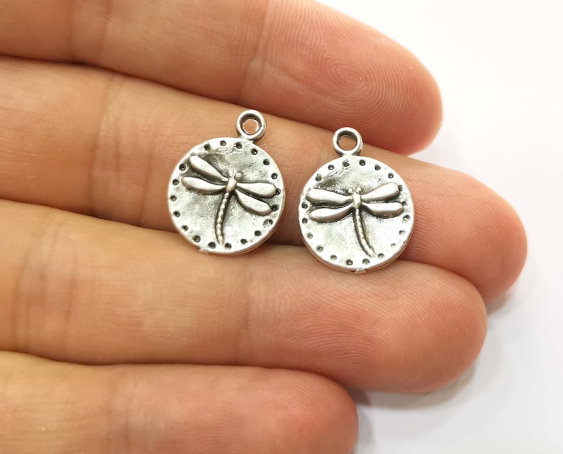 May include: Two silver-toned metal charms in the shape of a circle with a dragonfly design. The dragonfly is embossed on the charm and has a detailed design.