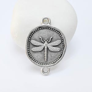 May include: A silver-toned metal charm with a dragonfly design. The charm is round with a beaded border and two loops for attaching to jewelry. The dragonfly is in the center, with detailed wings and body.