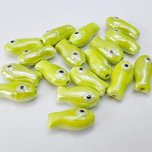 May include: A collection of small, glossy, chartreuse-colored ceramic fish beads. Each bead features a black eye and a rounded shape, perfect for jewelry making or craft projects. The beads are arranged on a white surface.