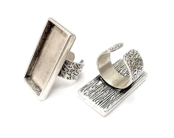 May include: Two silver-toned adjustable rings with rectangular bezels. The bezels are blank and ready to be filled with resin or other materials. The rings have a textured design.