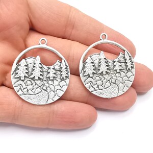 May include: Two silver metal charms in the shape of a circle with a cut-out design of a mountain range with trees. The charms have a textured surface.