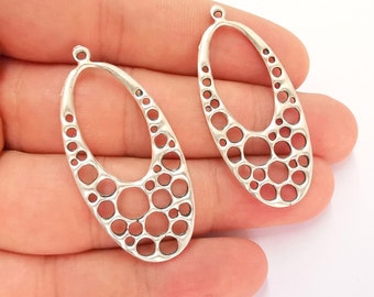 May include: Two silver-colored, open-work oval pendants. Each pendant has a small loop at the top and a series of varying-sized circular cutouts. The pendants are displayed against a white background.